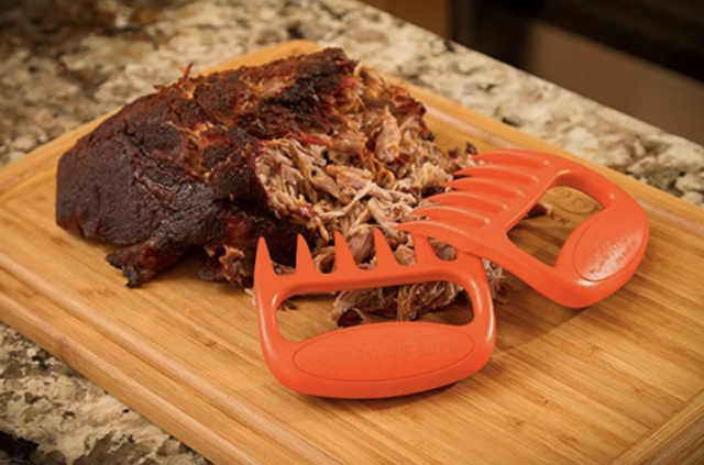 Large hunk of pork on bamboo cutting board with bear claw shredders laying on one end with shredded meat nearby. 