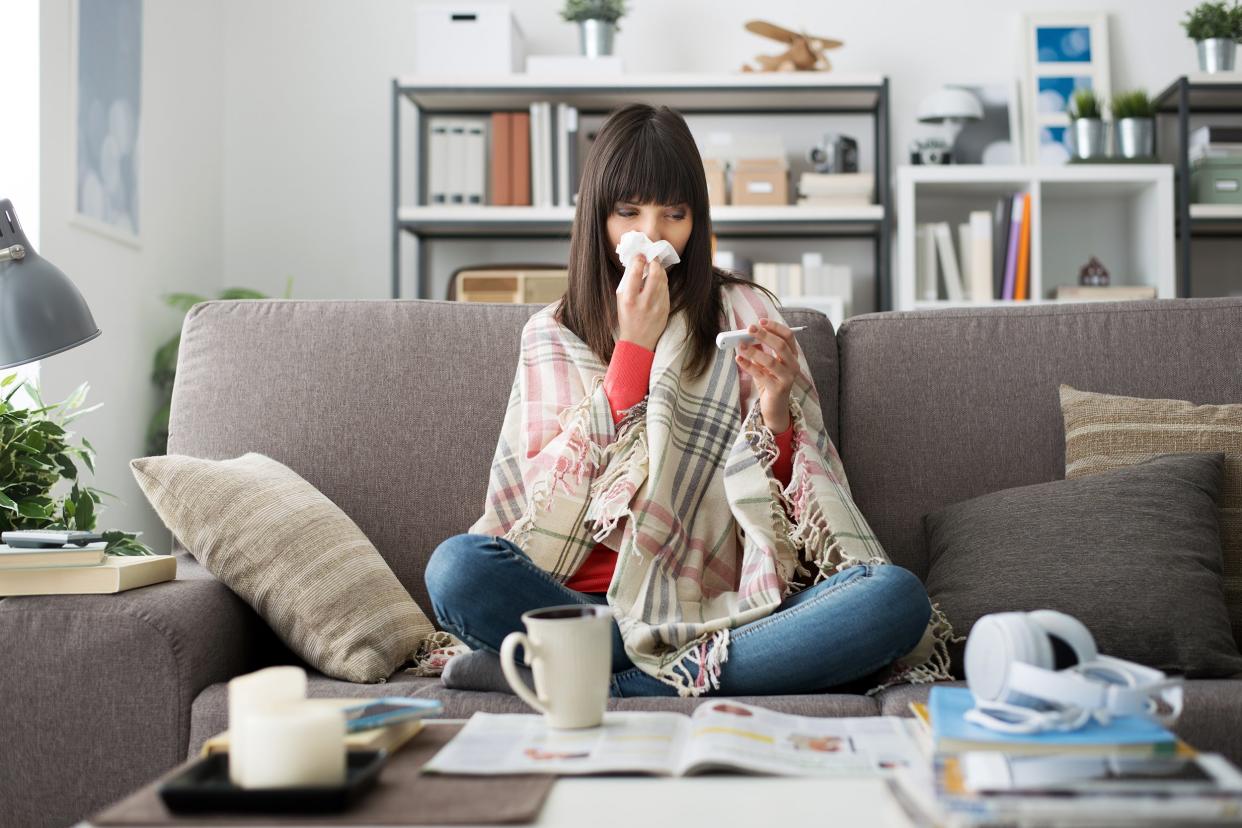 Sick woman with cold or flu sitting on the sofa at home taking her temperature