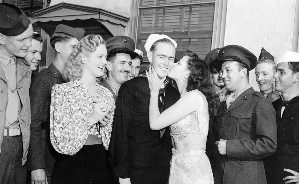 Yvonne De Carlo plants a resounding kiss on S1/C Harold Farnlund, Mountain Home, Idaho, as other servicemen wait their turn. Yvonne had to kiss the first 50 servicemen she met on Hollywood Boulevard and actress Martha O'Driscoll (Left) keeps tab of the payoff