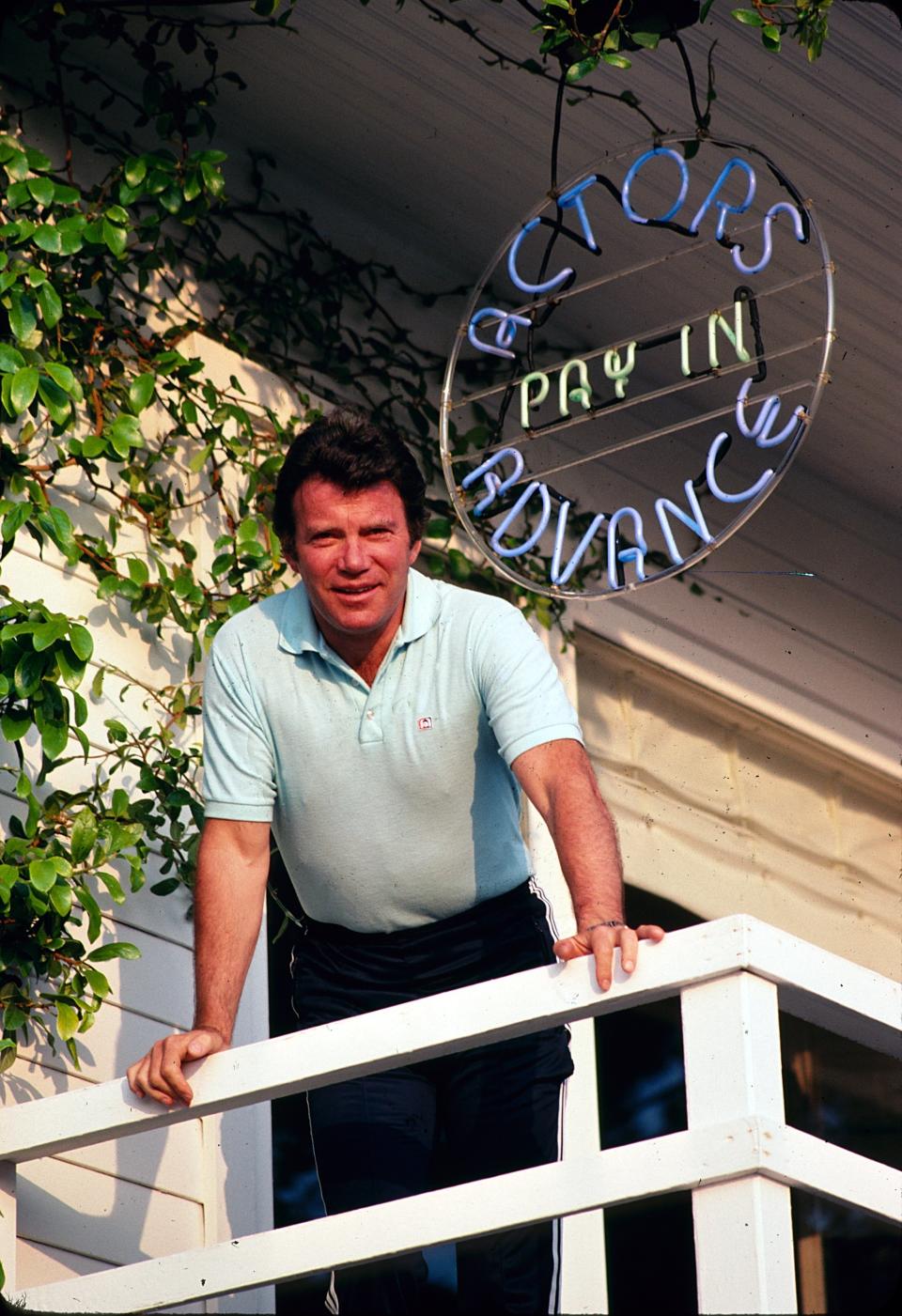 Man leaning on balcony rail below a neon sign that reads "ACTORS PAY IN ADVANCE."