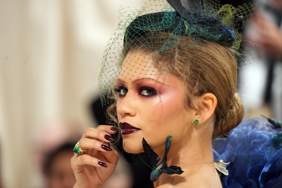 Zendaya with dramatic makeup and a veiled headpiece touches her face lightly