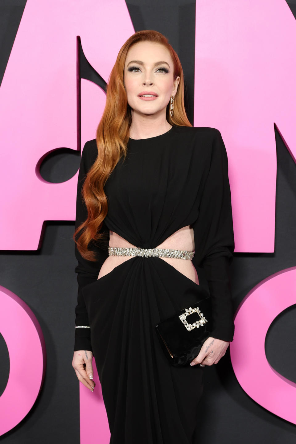 Lindsay Lohan posing on the red carpet for the premiere of Mean Girls wearing a black Alexandre Vauthier cutout dress, Roger Vivier clutch bag, and Giuseppe Zanotti strappy sandals.