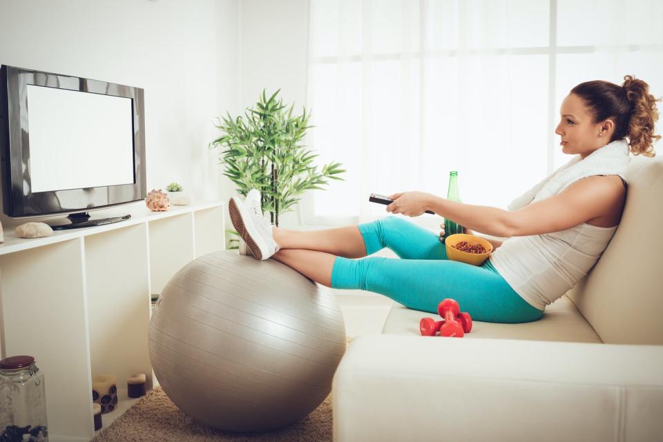 A woman in fitness clothes sitting on a sofa snacking and ignoring her fitness equipment while turning on the tv
