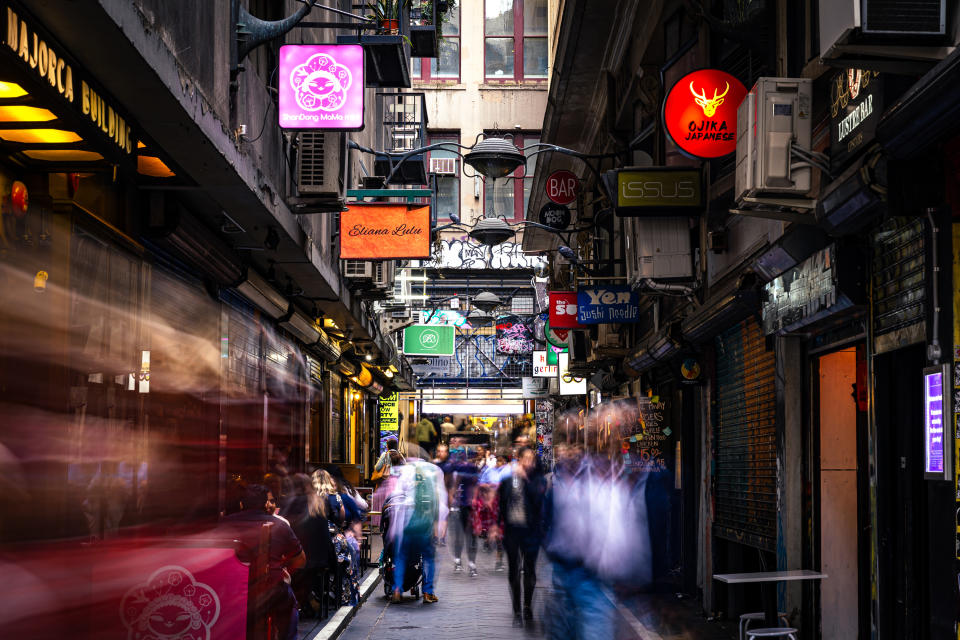 Crowded narrow alley with blurred moving people, surrounded by numerous shops and neon signs. Urban nightlife scene