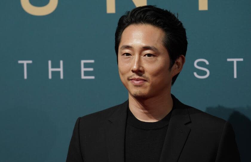 Steven Yeun is posing with a slight smile, his mouth closed, while wearing a black suit jacket and black t-shirt