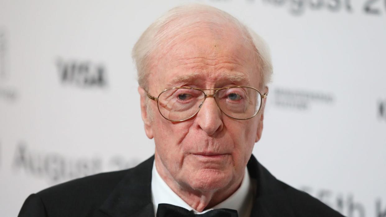 michael caine stands in front of a white blurred background and wears a black suit jacket, black bowtie, white collared shirt and large wire rimmed glasses, he looks to the left of the camera with a solemn expression on his face
