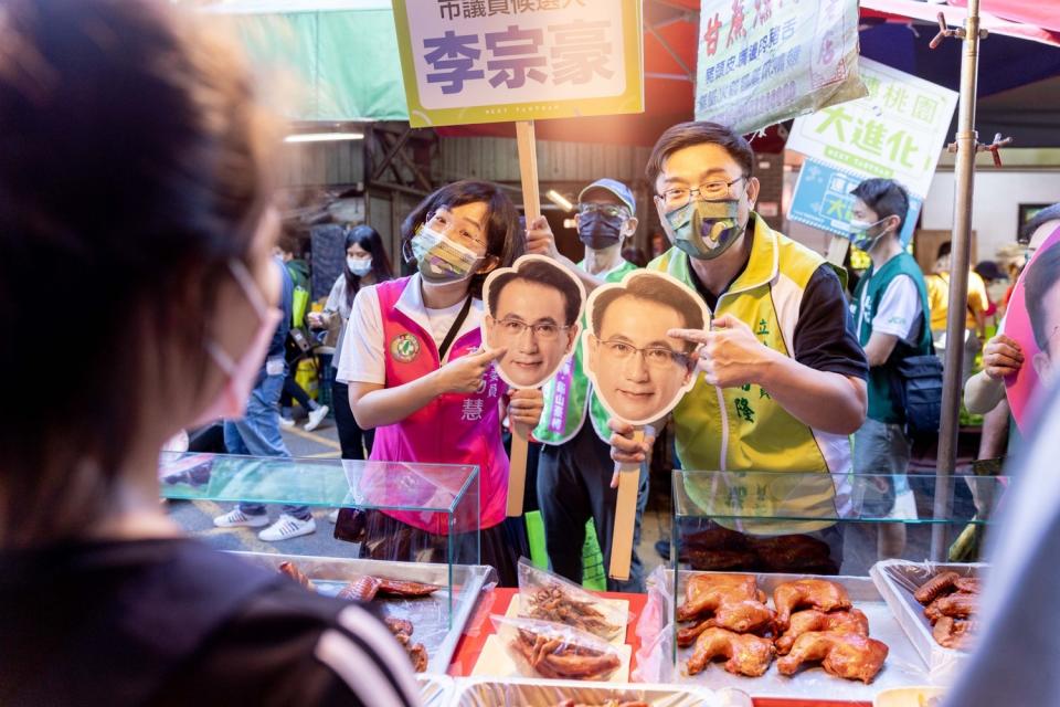 蘇巧慧及賴瑞隆手持鄭運鵬面具化為分身，在龜山市場掃街向攤商民眾拜票。   圖：鄭運鵬競辦／提供