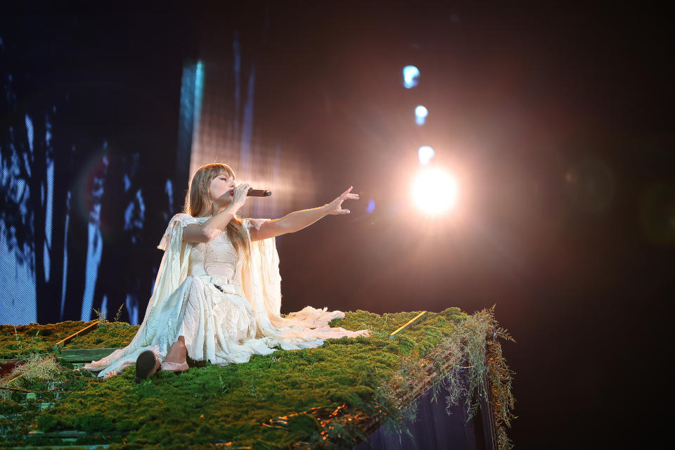 Taylor Swift in a white flowing dress sings onstage with a spotlight in the background