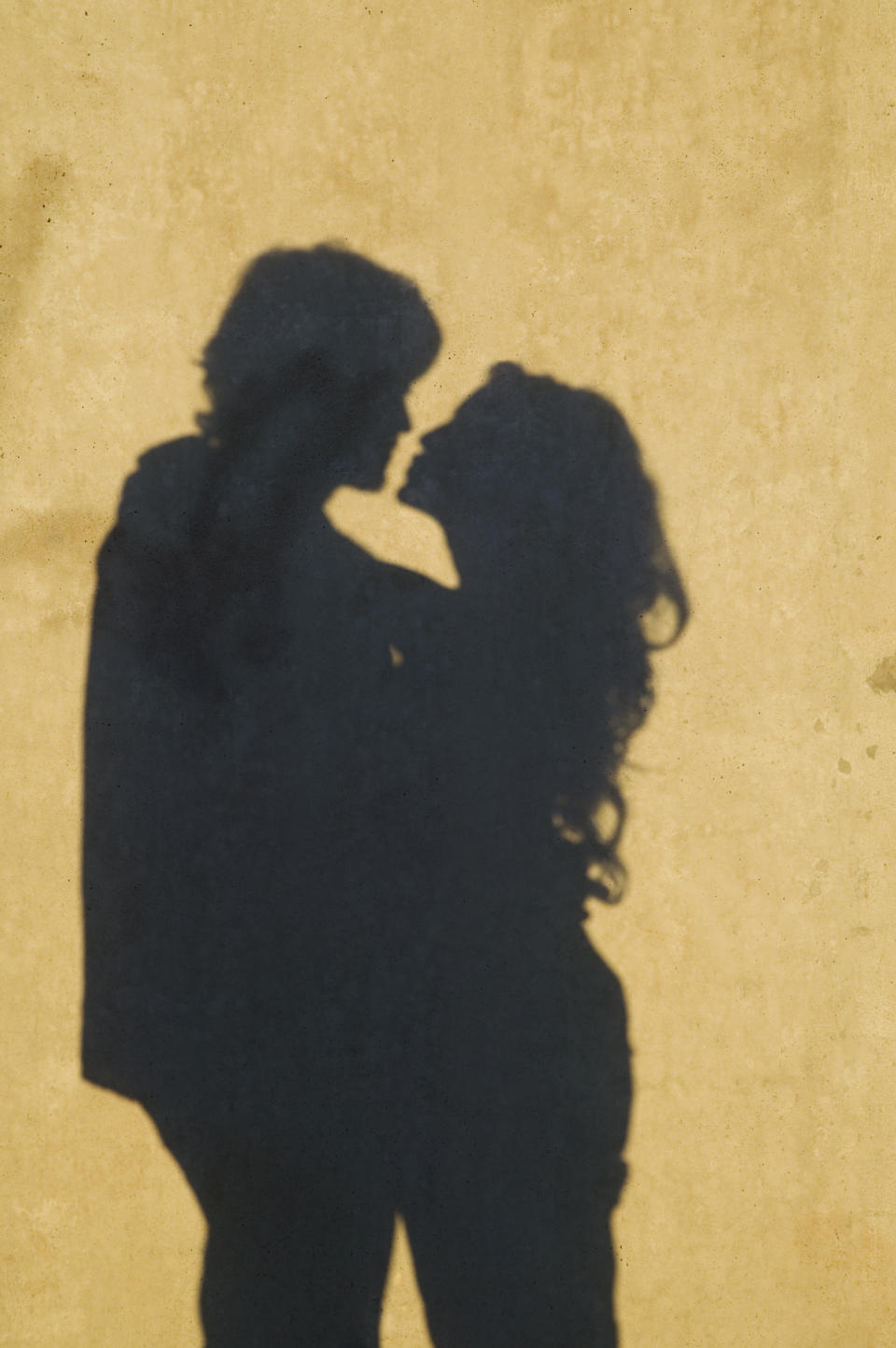 Shadowy silhouettes of a couple, facing each other intimately with heads close, suggestive of a romantic moment. The image exudes themes of love and connection