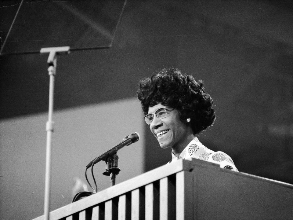 Black and white photo of Shirley Chisholm smiling and speaking at a podium.