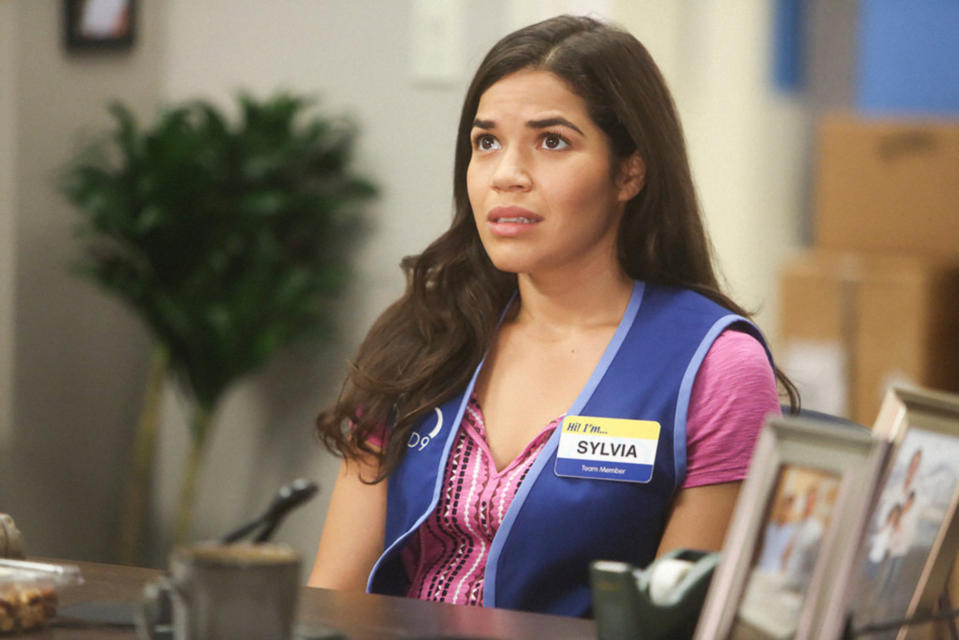 America Ferrera wearing a store employee vest, sits at a desk and looks ahead with a concerned expression