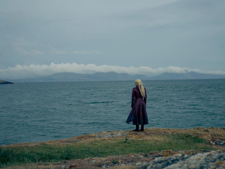 emma d'arcy as rhaenyra targaryen, seen from the back. rhaenyra's wearing a red lather coat, her hair loose, and she's seen from the back as she looks out to the se