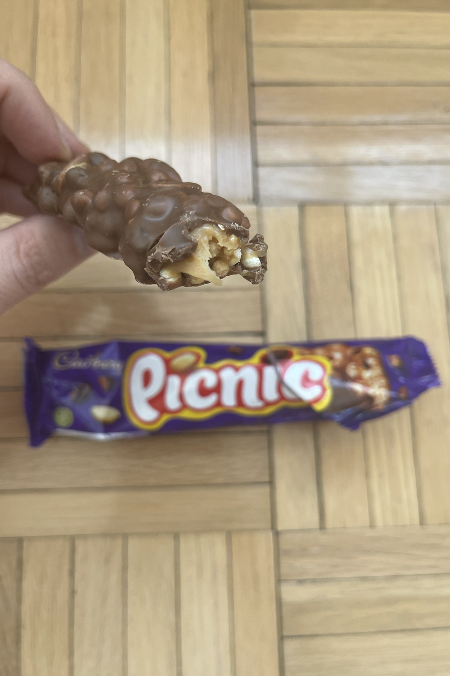 Hand holding a half-eaten Picnic chocolate bar over a wrapped one on a wooden floor