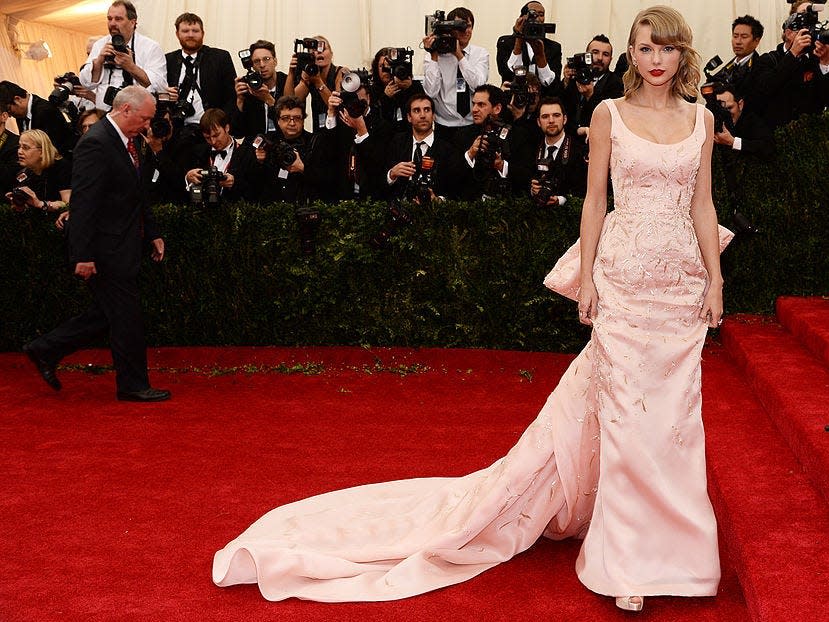 Taylor Swift poses at the bottom of the Met Gala stairs on the red carpet, wearing a pink gown with a long train.