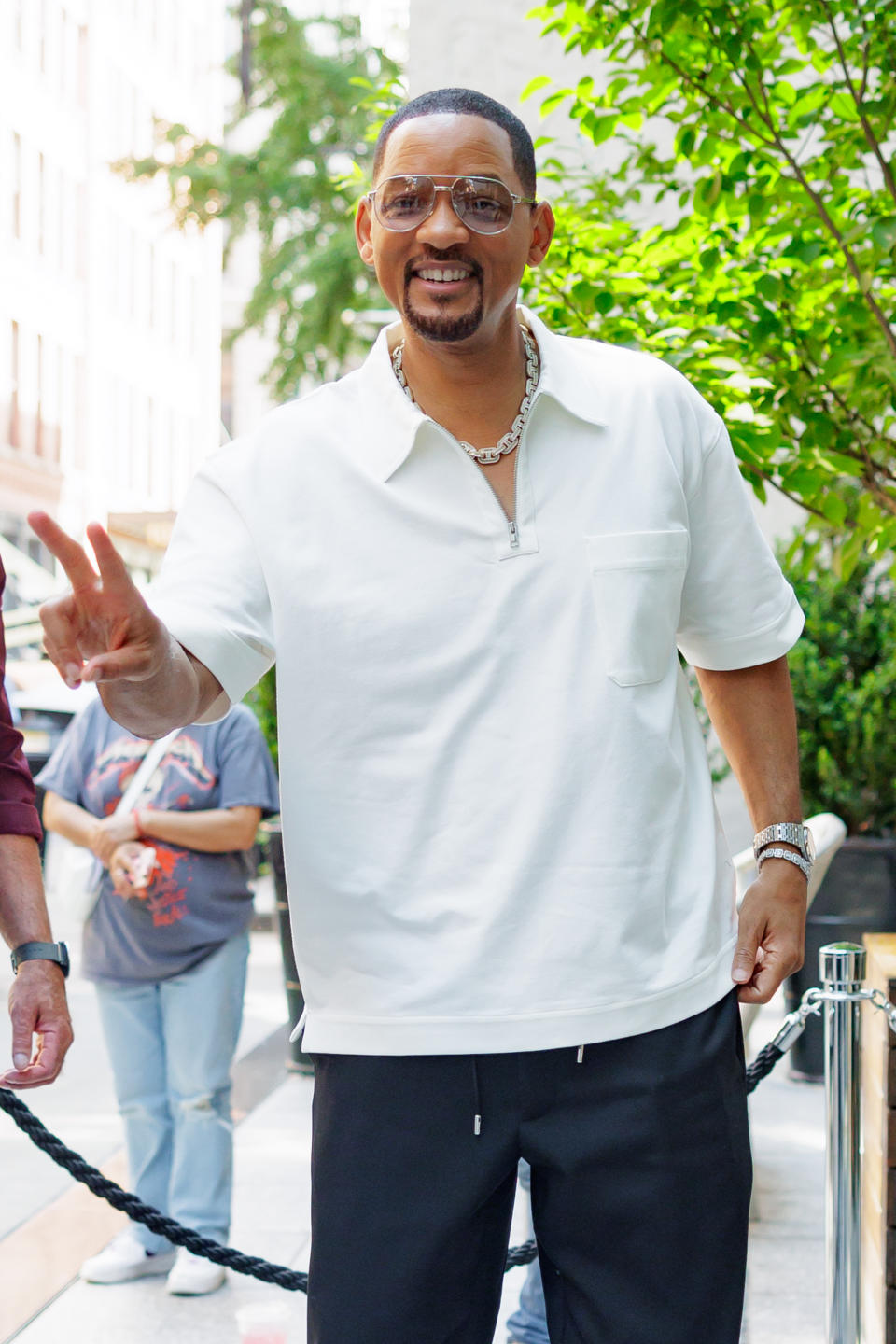 A man in casual attire, wearing a white shirt and black pants, gives a peace sign outdoors