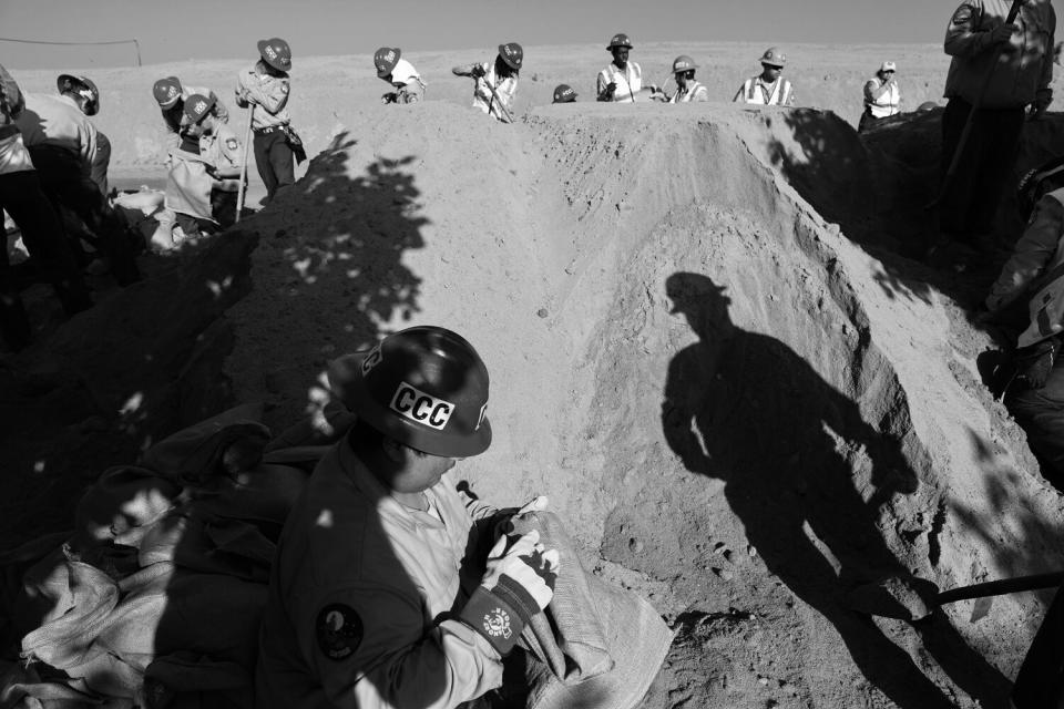 Workers, some with shovels and hard hats marked CCC, stand around a pile of sand.