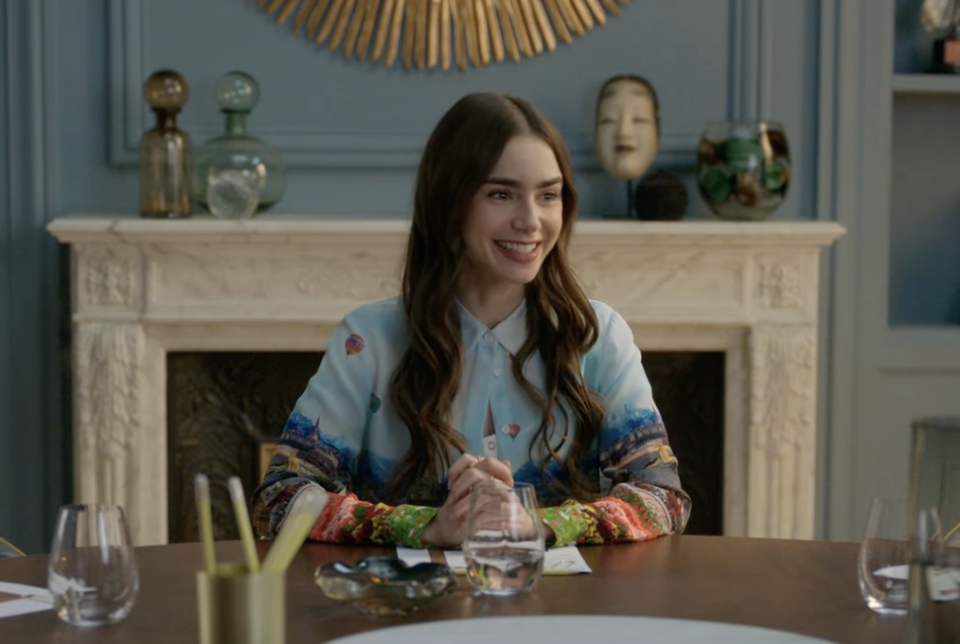 Lily Collins sits smiling at a round table in a stylish room with a fireplace and various art pieces