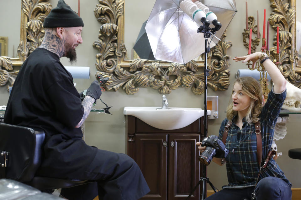 In this Thursday, April 11, 2019 photo Berkshire Eagle photographer Stephanie Zollshan, right, speaks with tattoo artist Brian Brown, of Dalton, Mass., left, at his tattoo parlor, in Dalton. Zollshan photographed Brown for a Berkshire Eagle section called Berkshire Landscapes. The paper now features a new 12-page lifestyle section for Sunday editions, a reconstituted editorial board, a new monthly magazine, and the newspaper print edition is wider. That level of expansion is stunning in an era where U.S. newspaper newsroom employment has shrunk by nearly half over the past 15 years. (AP Photo/Steven Senne)