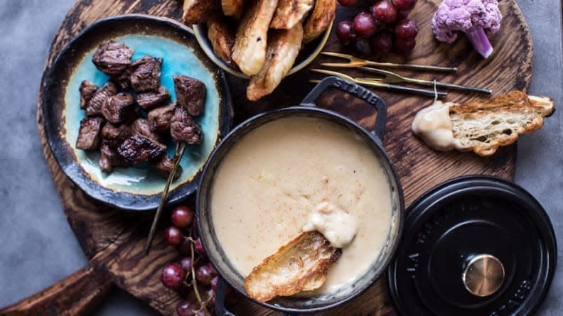 A bowl of gooey cheese fondue on a wooden cutting board with accompaniments