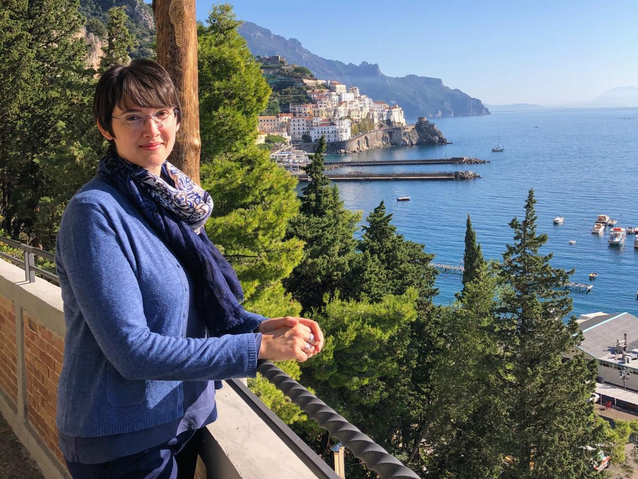 Laura poses at an overlook on the Amalfi Coast. There are buildings in the mountains in the background and boats in the water.