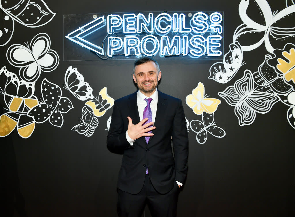 Entrepreneur Gary Vaynerchuk in a black suit with a white shirt and purple tie standing in front of a neon sign that says "Pencils of Promise"