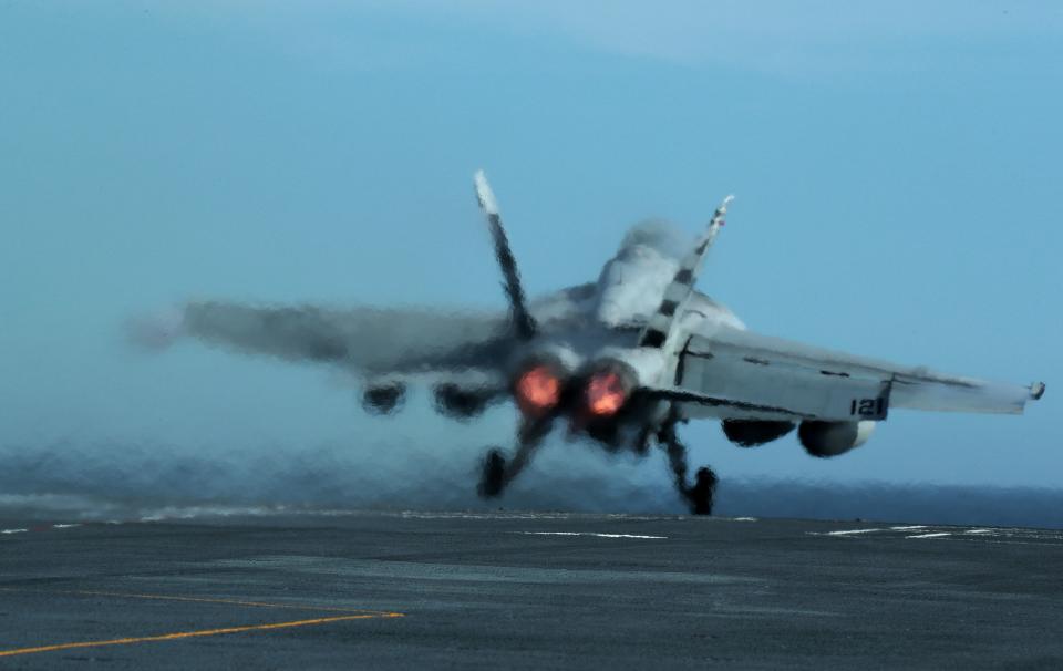 An American jet takes off from an aircraft carrier, a faint smear of jet fuel trailing behind it.