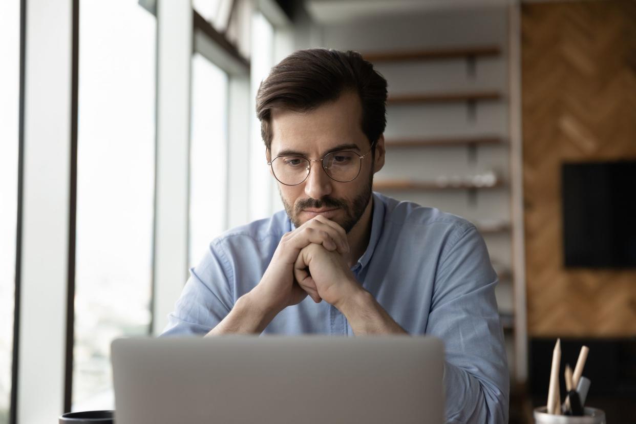 Thoughtful focused business man in glasses working at laptop computer, reading on screen, thinking, looking at monitor with serious face. Adult student watching learning webinar. Distance studying