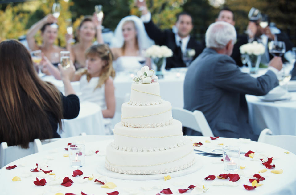 Wedding reception scene with a four-tiered cake in the foreground. Wedding guests in formal attire sit around tables and raise toasts to the bride and groom in the background