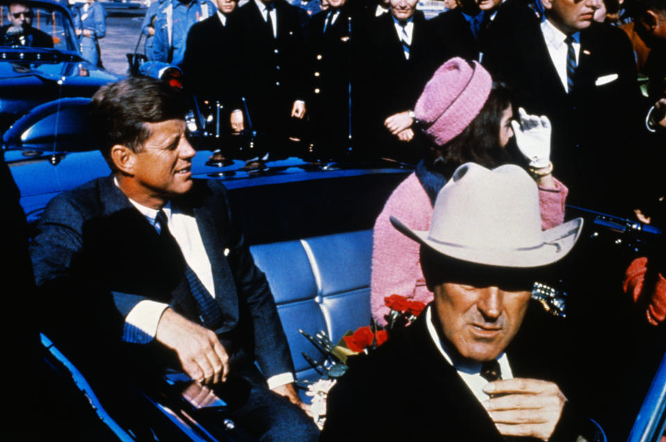 John F. Kennedy, Jacqueline Kennedy in pink suit and hat, and Governor John Connally in a car during a motorcade, with officials nearby