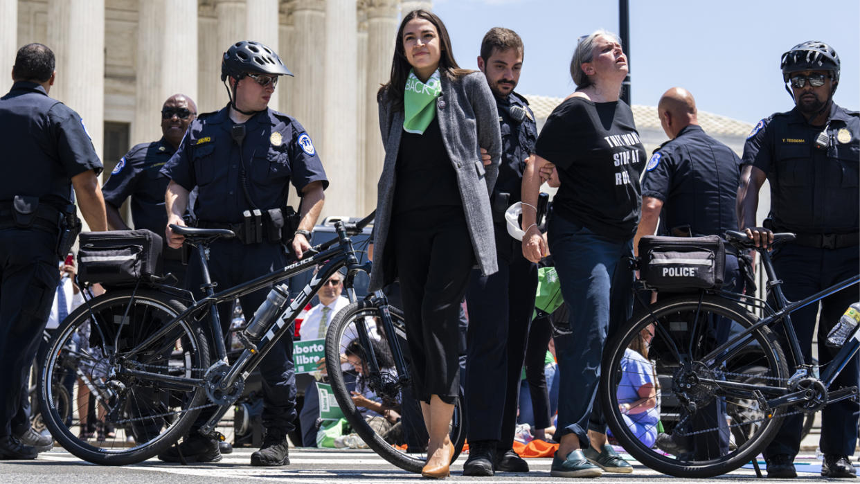 Rep. Alexandria Ocasio-Cortez, D-N.Y., center, looking upbeat, with one hand pulled behind her back by an officer, who holds the arm of another protester who looks thoroughly upset. Five other officers on bicycles stand by.