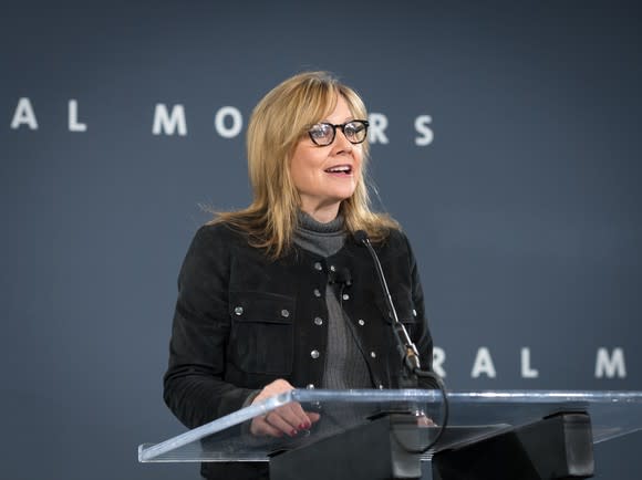 Mary Barra speaking at a podium in front of a backdrop with General Motors' logo.