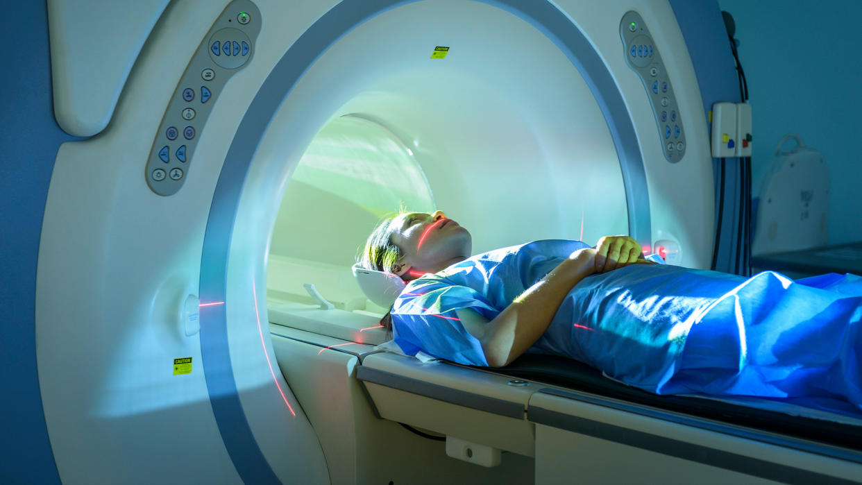  White woman in a blue surgical gown laying down as she's sliding into an MRI scanner. 