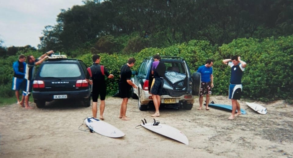 A group of men near two parked station wagons get ready to go surfing.