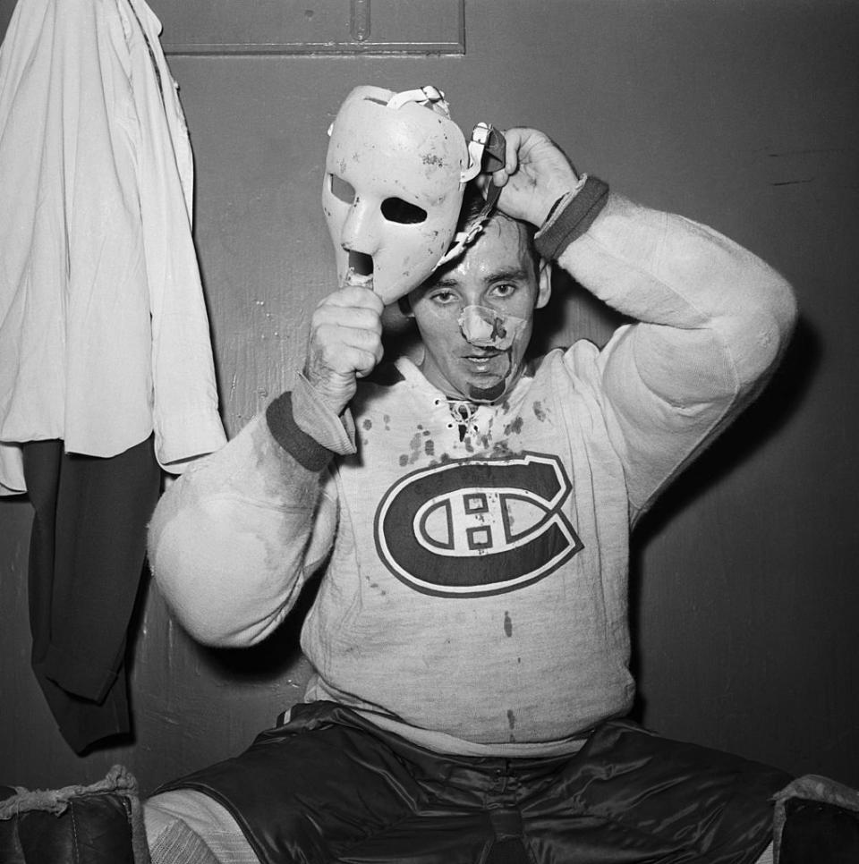 Jacques Plante, wearing a Montreal Canadiens jersey, sits in a locker room holding a hockey mask. His face shows signs of injury