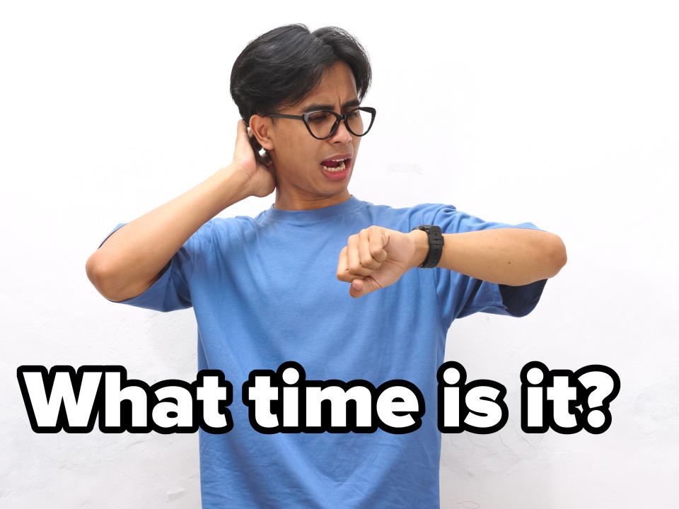 A man wearing glasses and a plain t-shirt looks at his watch with a worried expression, scratching his head