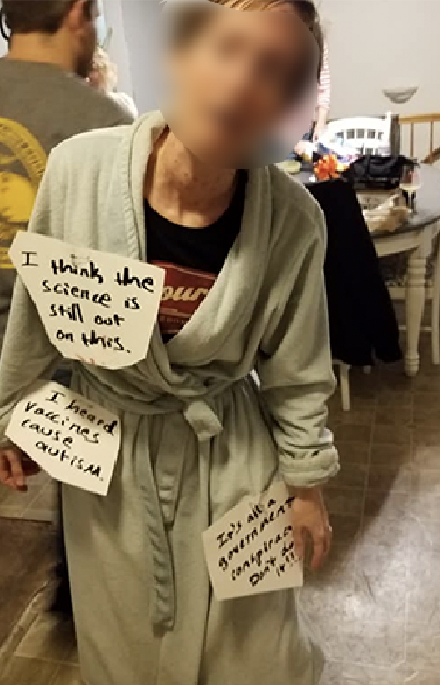 woman in anti-vaxxer costume with a bathrobe and signs saying "I heard vaccines cause autism" and other anti-vaxxer sentiments