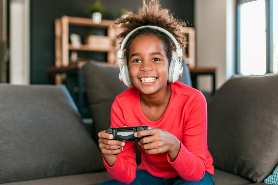 A young girl smiles while playing a video game, wearing a long-sleeved shirt and headphones, seated on a couch