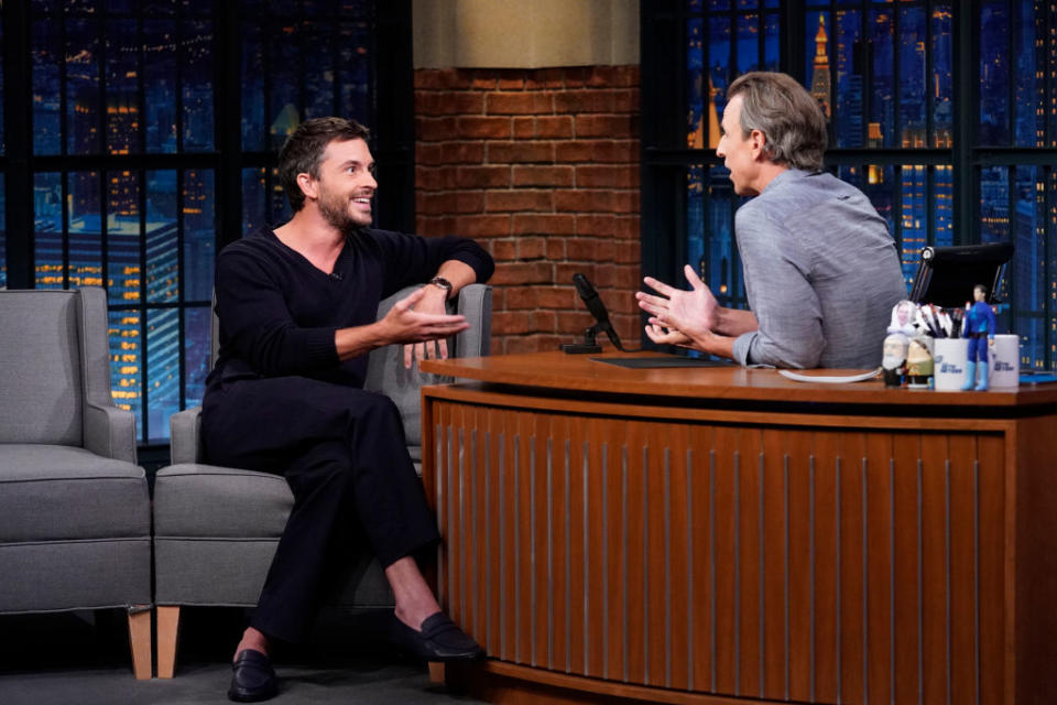 Jonathan Bailey, dressed casually, sits on a talk show set, engaged in conversation with Seth Meyers, wearing a button-up shirt