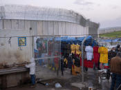 A vendor sells goods to Palestinian laborers as they cross from Israel back to the West Bank at the end of working day, next to a section of Israel's separation barrier in Meitar crossing in the West Bank, Thursday, March 10, 2022. Twenty years after Israel decided to built its controversial separation barrier amid a wave of Palestinian attacks, it remains in place, even as Israel encourages its own citizens to settle on both sides and admits tens of thousands of Palestinian laborers. (AP Photo/Oded Balilty)