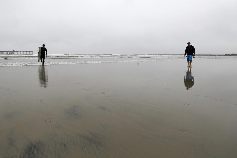 A man wears a mask while walking on a beach as a surfer makes his way out to the waves near the Ocean Beach pier Thursday, April 30, 2020, in San Diego. A memo sent to California's police chiefs says Gov. Gavin Newsom intends to close all beaches and state parks starting Friday in the wake of a weekend that saw a crush of people at open seashores. (AP Photo/Gregory Bull)