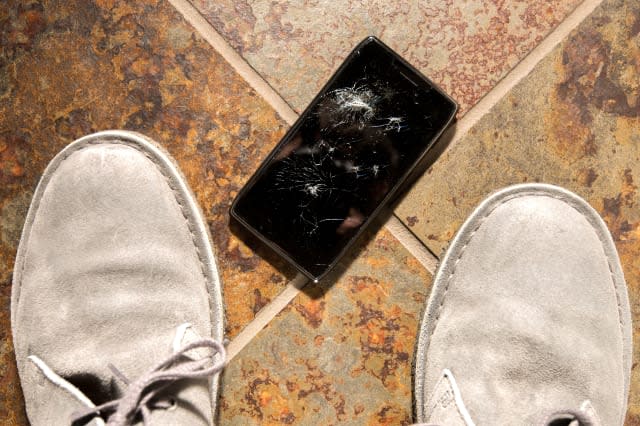 A smartphone lies broken between the shoes of its owner just after being dropped.