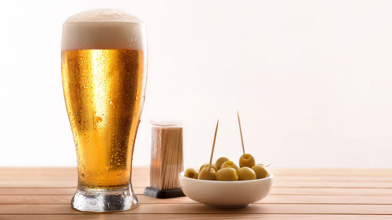 Beer in pint glass beside bowl of olives and toothpick holder