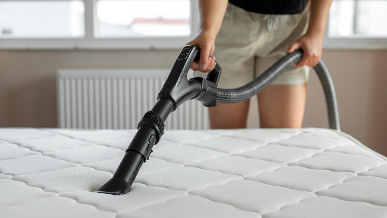  Woman wearing beige shorts vacuuming her white mattress to get rid of bed bugs. 