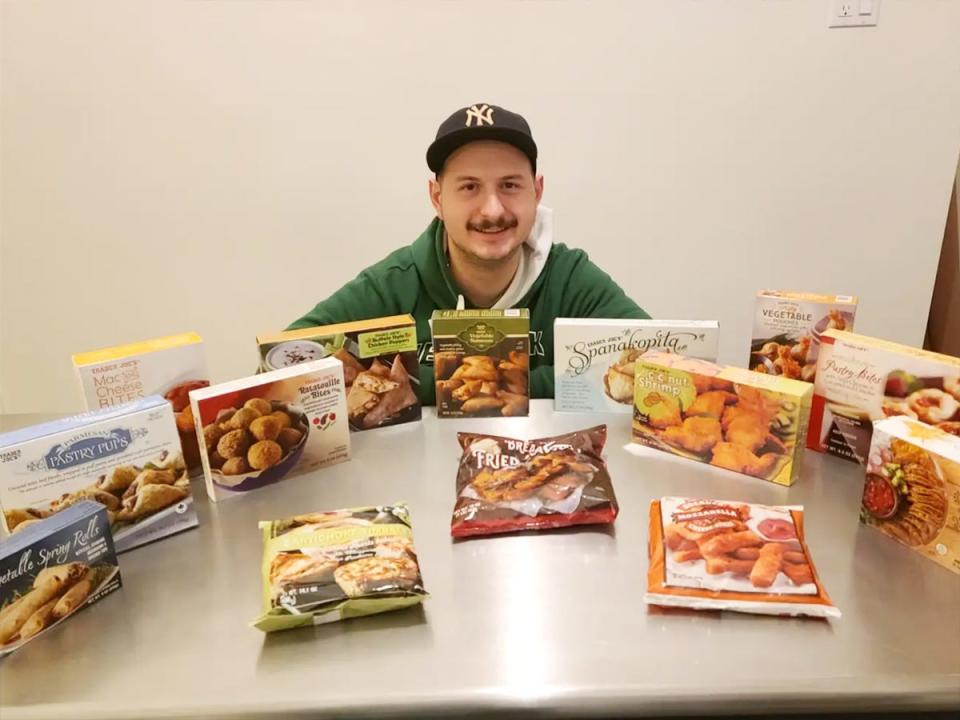 The writer, wearing a green hoodie and black baseball cap, sits at a table full of Trader Joe's appetizers