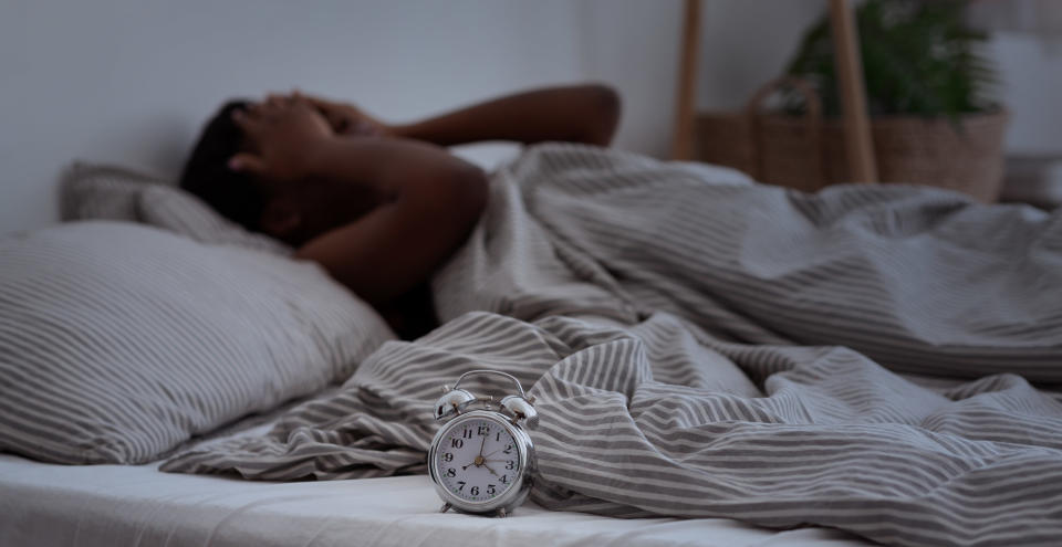 A woman laying in bed with her hands to her head and an alarm clock in the bed with her