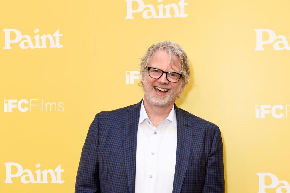 brit mcadams, wearing a blue patterned suit jacket and glasses, smiles directly into the camera, with a yellow wall behind him