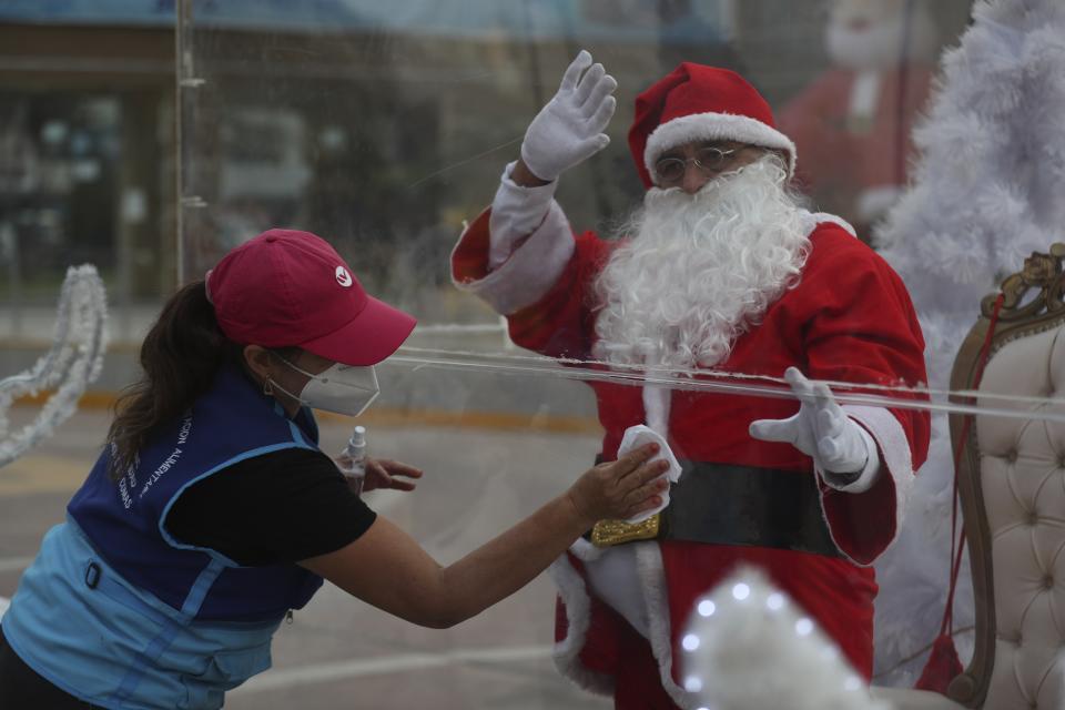 Dressed as Santa Claus, David Pizarro waves from inside a plastic enclosure as a woman disinfects it in an effort to curb the spread of COVID-19, at a square in the Comas municipality of Lima, Peru, Monday, Dec. 21, 2020. (AP Photo/Martin Mejia)