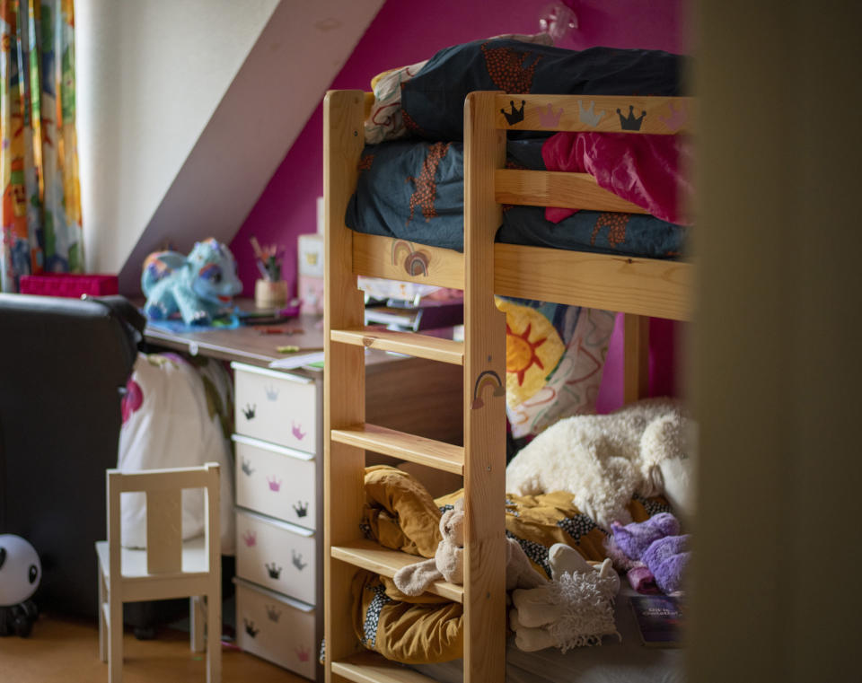 A cozy child's bedroom with a loft bed, stuffed animals, and colorful decorations. A desk with art supplies and toys is next to the bed