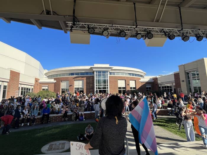 Students crowd outside a school building; one is holding a trans pride flag