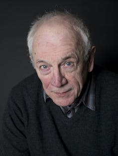 An elderly bald headed man in a dark shirt and jumper.